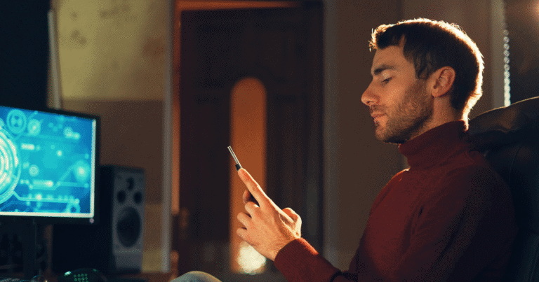 A man is sitting in front of a computer while looking at his mobile phone
